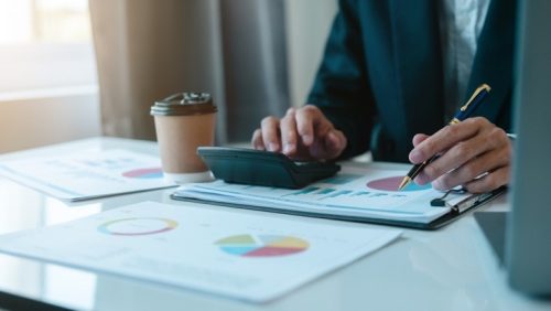 Stock photo, printouts of charts and graphs on a table, with one person writing on paper and using a calculator