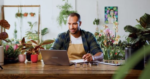 Stock photo, man sitting at a table inside a plant shop, looking at a laptop and writing on a piece of paper