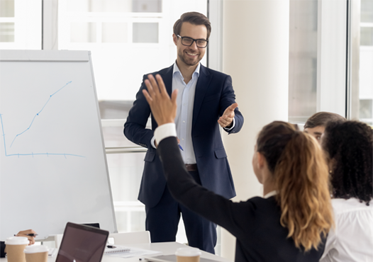 Stock photo, man giving presentation in a conference room to a group of professionals, one woman at the table raising her hand